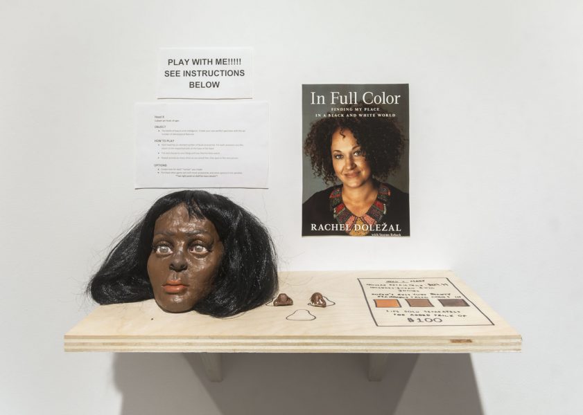 Interactive installation on the wall there is a sign that says "Play with me!!! See instructions below" in black capital text, below it are a set of instructions, next to this on the right is an image of a black woman with natural curly hair and there is a title that says "In Full Color" in white text, installed on the wall is a wooden shelf that has a mask with black hair and dark skin, two fake noses, and some drawings on the wooden shelf in black lines