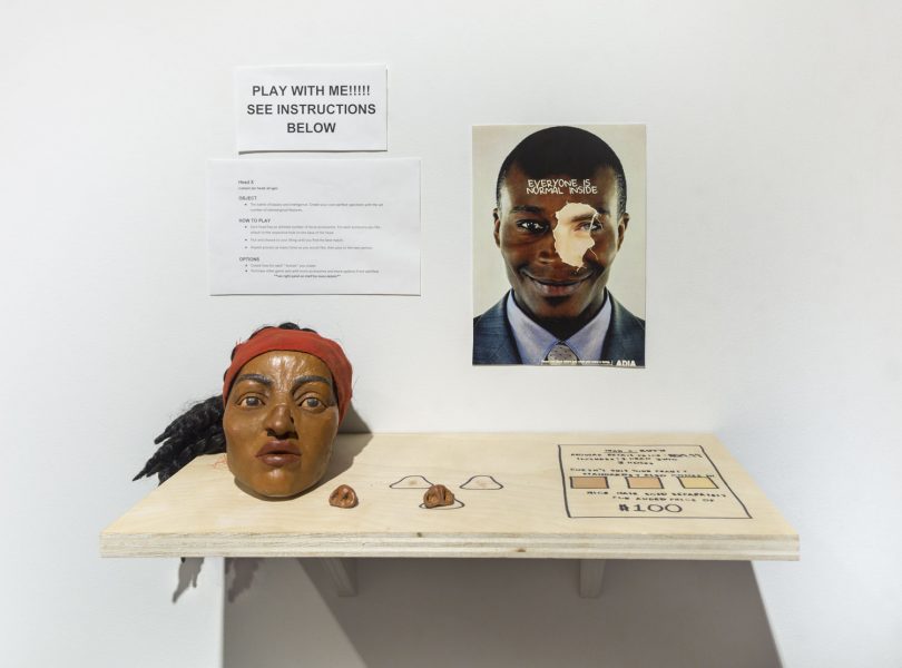 Interactive installation on the wall there is a sign that says "Play with me!!! See instructions below" in black capital text, below it are a set of instructions, next to this on the right is an image of a black man with a snippet of a white person's eye over the right eye of the black man, installed on the wall is a wooden shelf that has a mask with black hair, a red headband, and dark skin, two fake noses, and some drawings on the wooden shelf in black lines