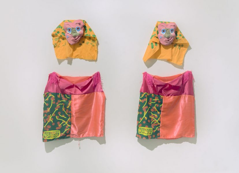 Two wall sculptures hung up next to each other, they both are masks with pink faces, blue eyes, and green noses, on a yellow piece of fabric with green polka dots, below each one there is a shirt that is sewn with pink fabric on top, and green fabric with neon yellow lines and accents, orange dots, and peach fabric next to it