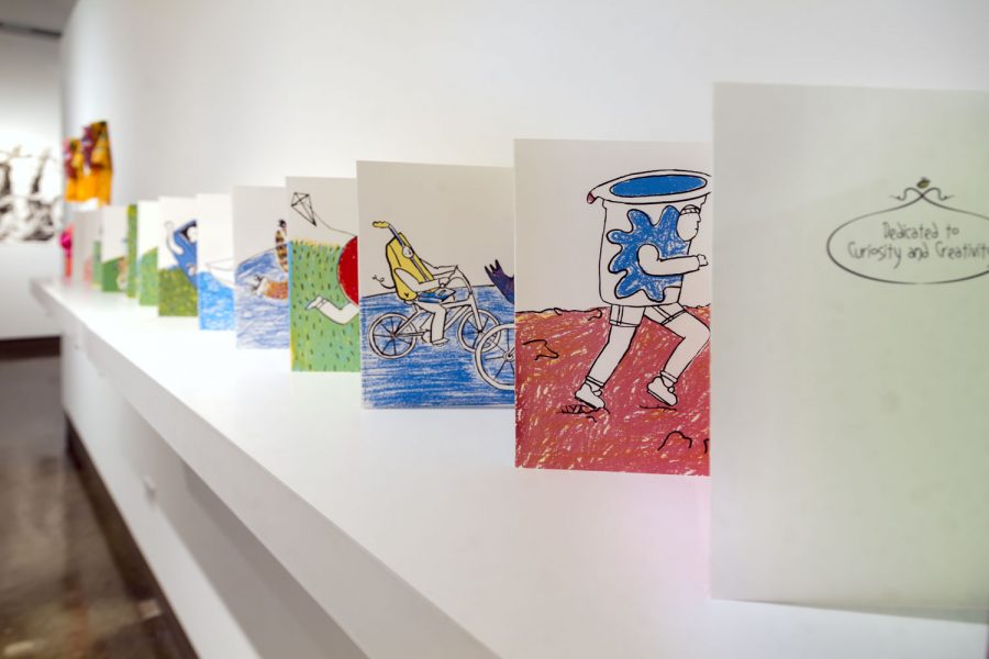 Installation view of a white wall shelf with several rectangular drawings propped up next to each other there is a cup with human legs running on red dirt, a banana riding a bike, and other similar illustrations that continue