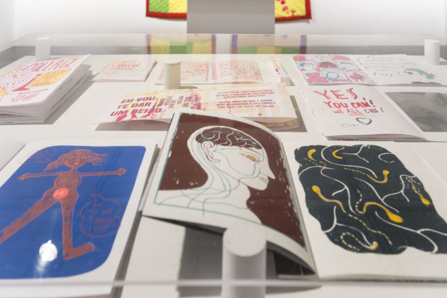 Close up of various books and prints on a table, the closest one has a red figure of a woman on a dark blue background, a portrait side view of a person showing their brain on a maroon background and an abstract illustration on a black background with white and yellow lines