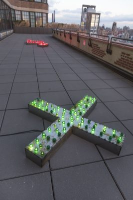 A sculpture of a metal X with green round edison lightbulbs on top of it, some of the lightbulbs are dim and not lit up but most of them are, in the distance there is a similar piece shaped like an arrow with red lightbulbs