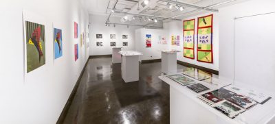 Various prints are hung on the wall on the left and back of the gallery, on the right wall there are two yellow and green checkerboard quilted textile hangings with a red border and purple and white silkscreen piece in the center, there are pedestals that display various books and prints in the middle of the gallery