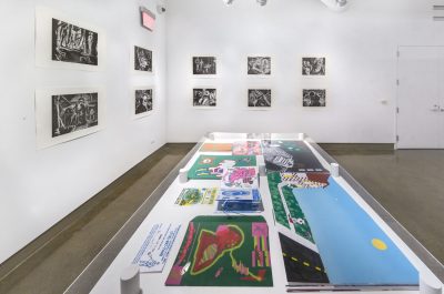 Gallery view with a table displaying various books and prints in the foreground, in the back there are ten black and white prints/drawings hung up on the wall in two rows of five