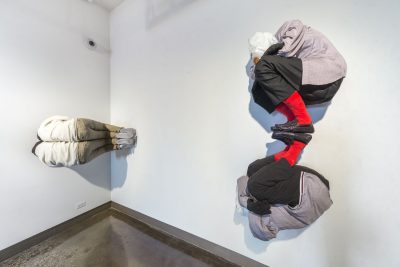 Sculpture of two people with their knees bent and face in one of their arms, there are plastic bags over their heads, and they are installed against the white wall connected at the feet so one is right side up and the other is upside down like a reflection of the first one, to the left there is a sculpture of two people right next to each other in the same pose, but stuck on the side and facing the gallery wall so you can't see their front