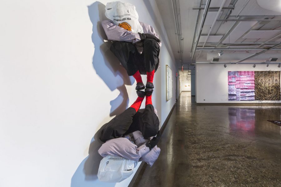 Sculpture of two people with their knees bent and face in one of their arms, there are plastic bags over their heads, and they are installed against the white wall connected at the feet so one is right side up and the other is upside down like a reflection of the first one, there is a purple and pink and brown piece of artwork displayed in the background