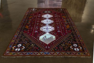 Red, black, white, blue, gold traditional styled rug is on the floor with some images and light projected onto the middle geometric motifs of the rug