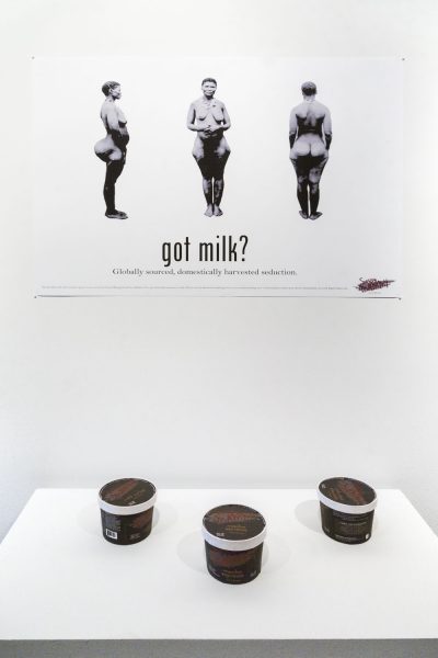 A print that has a side view, front view, and back view of a naked woman in black and white is printed with the words "got milk?" and a smaller caption typed out below it, in front of the print below is a pedestal with three containers that are black, white, with brown letters