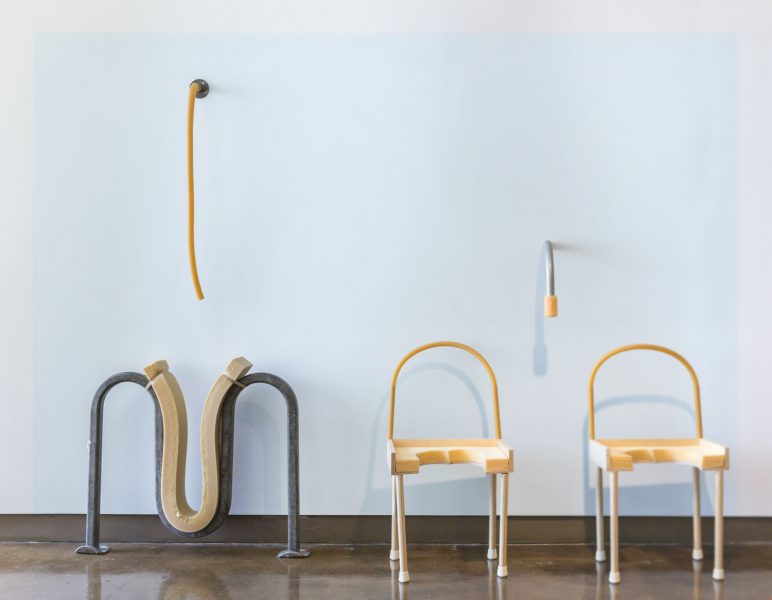 Installation with two tubes that are hanging down from the wall, on the floor there is a bike rack sculpture with a piece of foam on top of the middle bend, and two yellow chairs with no seats on the right of the bike rack