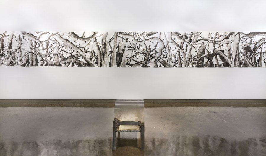 A black and white horizontal scroll drawing is hung up on the white wall, in the center on the floor there is a sculpture that looks like a stool or small table with a translucent case on top