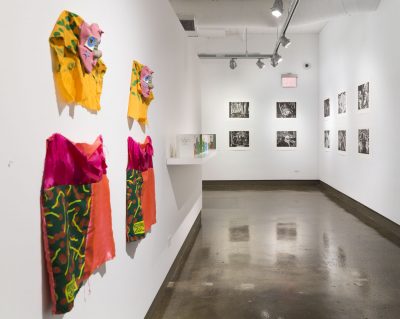 Four textile pieces are hung up on the wall on the left, in the background there is a wall shelf and a glass case displaying other pieces, there are ten black and white photographs hung up on the wall in the back and to the right