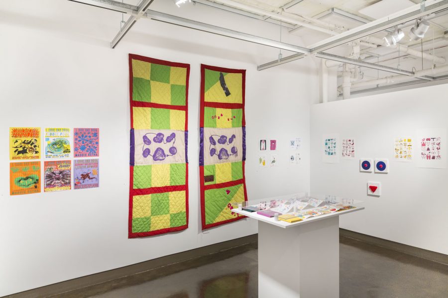 Gallery view with various colorful prints and drawings pinned on the wall, there are two long rectangular quilted textile hangings in the middle left wall with a red border, purple and white silkscreen, and yellow and green checkerboard pattern, in the center there is a white pedestal with books and other pieces on it