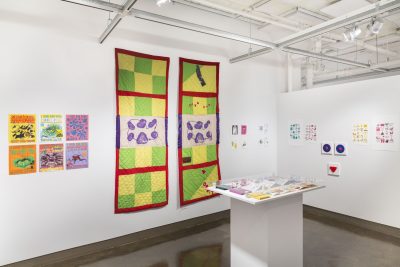 Galery view with various colorful prints and drawings pinned on the wall, there are two long rectangular quilted textile hangings in the middle left wall with a red border, purple and white silkscreen, and yellow and green checkerboard pattern, in the center there is a white pedestal with books and other pieces on it