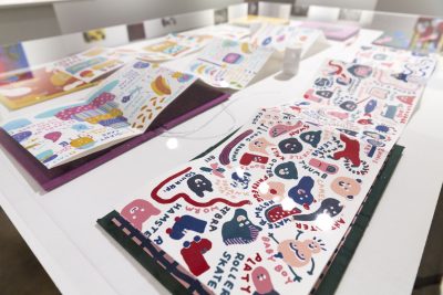 Close up of some books laid out on a white table, the books are colorful with various illustrations in them, the closest one has illustrations of people and animals with text surrounding each image