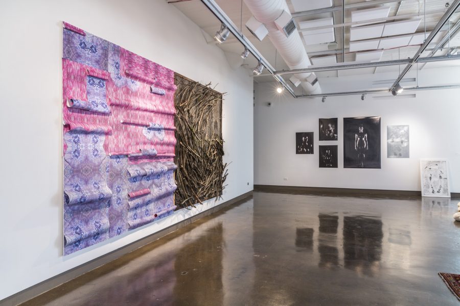 A purple and pink tapestry piece next to a brown art piece with branches on the left white wall, five black and white photographs of various sizes on the back wall, a black and white print leaning on the back wall on the floor to the right