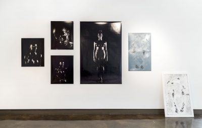 Five photographs hung on the wall, from left to right there are three smaller black and white photographs of a person, a large rectangular photograph of the same person, a smaller light blue photograph, and one photograph that is on the floor leaning against the wall