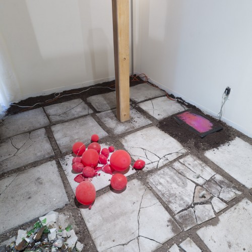 A floor made of stone slabs on top of fine soil with shattered glass on it, red balloons, and a screen partially covered with soil