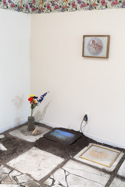 A floor made of stone slabs with flowers in a recipient, a screen partially covered with soil, a print on one stone slab, and a print hanging on the wall.