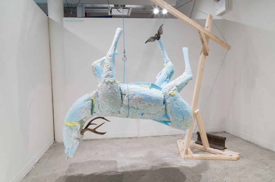 Installation view of an upside-down deer made of blue material. The deer is hanging from a wooden structure. Sculpture made by Vincent Chen