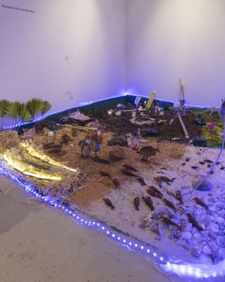 Installation with a bed of dirt, on top there is moss and some pebbles and flowers, a dinosaur, a pipe with a faucet attached, some mice, cigerette, a snow globe, and a toy truck, with a line of blue led lights on the outside around the installation, one part of the ground has hay, and the other side has sand and seashells as well as lobsters, fish, and various small figurines