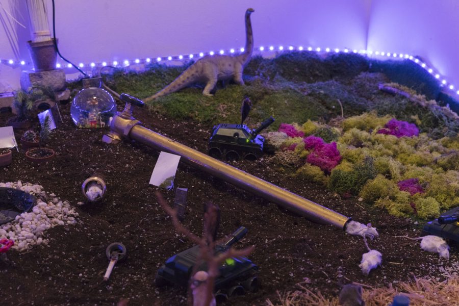 Installation with a bed of dirt, on top there is moss and some pebbles and flowers, a dinosaur, a pipe with a faucet attached, some mice, cigerette, a snow globe, and a toy truck, with a line of blue led lights on the outside around the installation