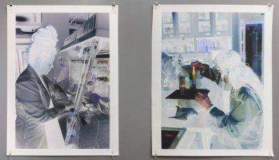 Two prints hung up on the wall with binder clips on all four corners, with a white border depicting negatives of people working in a laboratory, the one on the right shows a woman reaching into a machine, and the one on the right shows a woman measuring something at a table on a scale