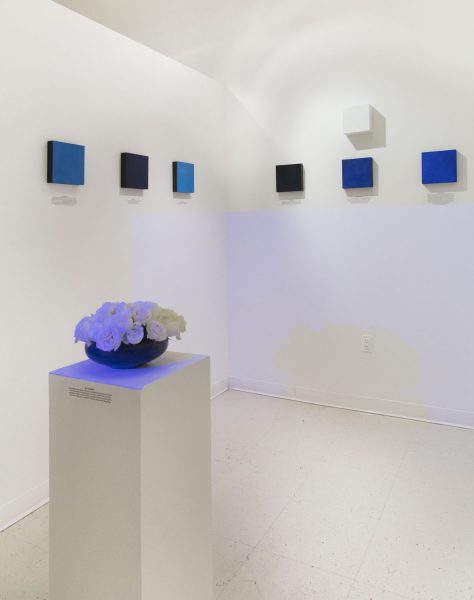 Six blue square paintings with one white square painting hung on the wall above the middle painting on the right, with a short wide vase containing white roses on a white pedestal in front with a black text caption printed below the paintings on the wall and also on the front of the pedestal