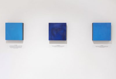 Three square blue paintings hung up with black text printed on the wall below the paintings, from left to right the paintings are light blue, dark blue, and medium blue