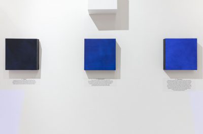 Four paintings installed on the wall with caption in black text printed on the wall below it, the paintings are square, from left to right they are painted dark blue, medium blue, bright blue, and the fourth painting is white and installed above the medium blue one