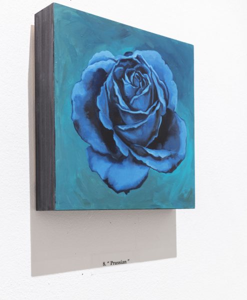 Square painting on a thick block of wood with gray sides hung up on the wall with black text below it printed on the wall that reads "& "Prussian" " the painting is a blue rose on a teal background