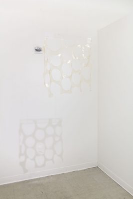 A clear sheet of plastic with circles cut out of it hung up in front of a white walled corner, with a light projected on it to cast a shadow