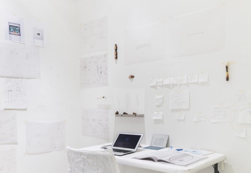 A chair and a desk wrapped in white with a laptop and some books on the desk, on the white wall around the desk are various white pieces of paper with notes and sketches as well as some brown specimen taped to the wall