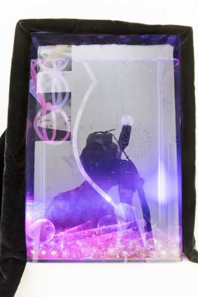 A silhouette of a person holding up a camera covered in layers of jewel like pieces on the bottom, and neon pink/purple lines drawn over with a black border