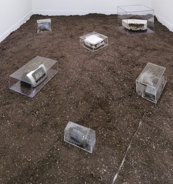 Six electronic devices inside clear acrylic boxes sitting on a bed of dirt