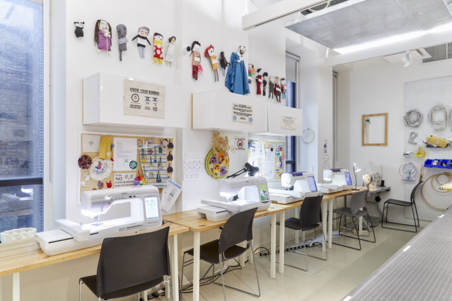 An interior view of the BFA Fine Arts Fibers Lab Facility, showing a row of digital embroidery stations with student artwork hanging above them.