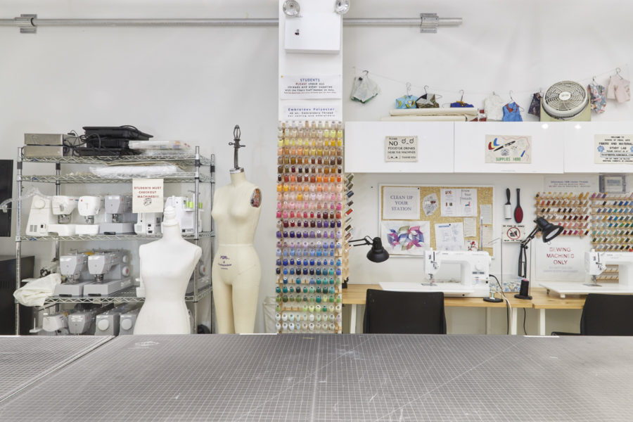 Fibers Lab Facility |SVA BFA Fine Arts. A large cutting table in the foreground with a rack of sewing machines and dress forms behind on the left and two embroidery machines on the right.