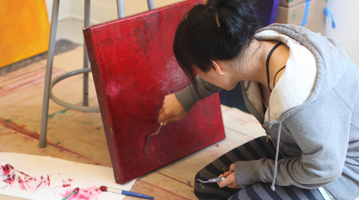 A student is working on her painting from a crouch position. The painting has the background in solid red color