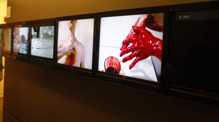 Installation of an array of TVs with images which contain a pair of hands with red paint on them, a person with color on chest, etc.