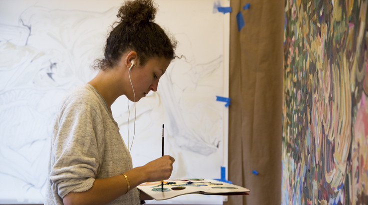 A student working on an abstract painting with her color sheet and the brush in her hand, listening to something in her headphones