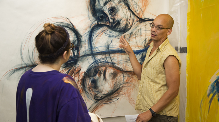 A person is poting his hand at a painting on the wall of two people made only of contour lines while he is talking to another person in front of him
