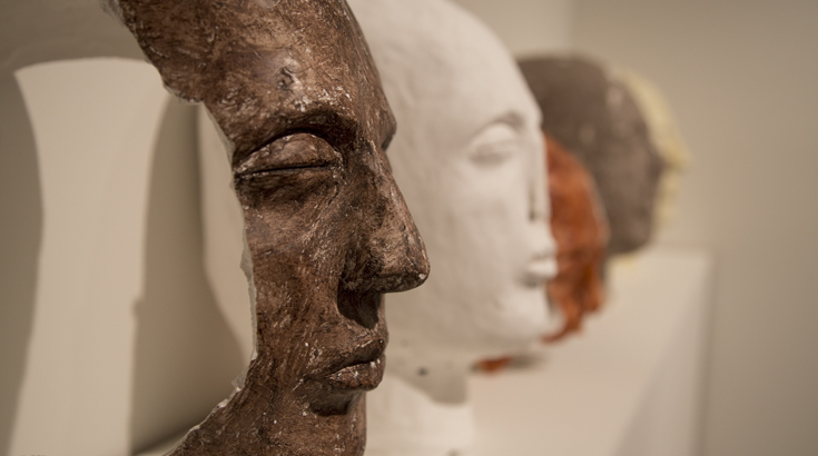 Side view of head sculptures made from different materials in bronze, white, orange, etc.