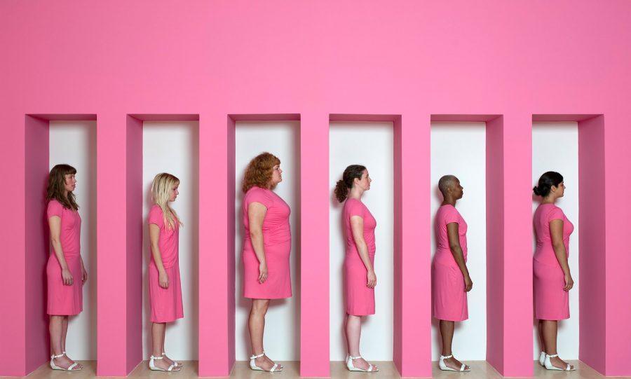 Image of six women of different ethnicities and ages dressed in mid-length pink dresses and white sandals. The six women are in a matching pink installation with slots they're standing in. All the women are facing towards the right, with their hands by their waists.