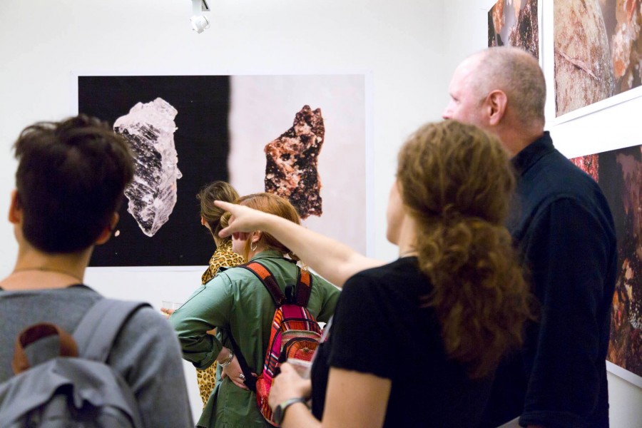 A few people take part in an exhibition with prints representing metals, crystals, and rock formation, and one of the men points to a specific print showing it to someone
