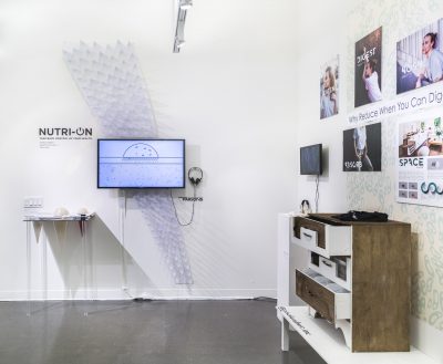 2017 BioDesign Challenge, installation view on the left there is a tv screen with headphones, a pedestal with sculptural pieces, to the right there are five photographs hung on the wall, with a smaller tv screen with headphones a pedestal with sculptures on top, and a raw wood and white dresser displayed next to it