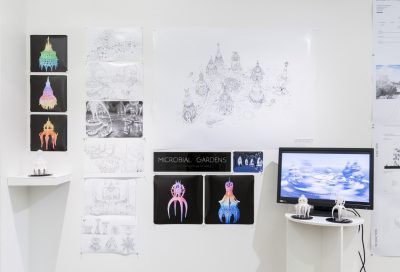 14 various drawings and diagrams of an alien like creature hung up on a white wall, there is a wall shelf to the left that holds a figurine that looks like the sketches on the wall, to the right there is a white pedestal with a tv screen and two alien figurines