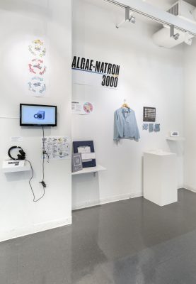 Installation shot of something called the Algae-Matron 3000, with a clue shirt hanging from the wall and a screen mounted on the wall with various posters.