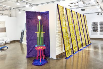 Six yellow metal structures with trim covered with purple faux fur. Installation has a giant module lamp structure with a bright pink base on top of faux fur carpet.