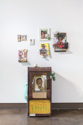 Salon style installation of mixed media collage items on wooden paintings. A sculpture is in front of the wall with another portrait, plastic bag, and deli coffee cup.
