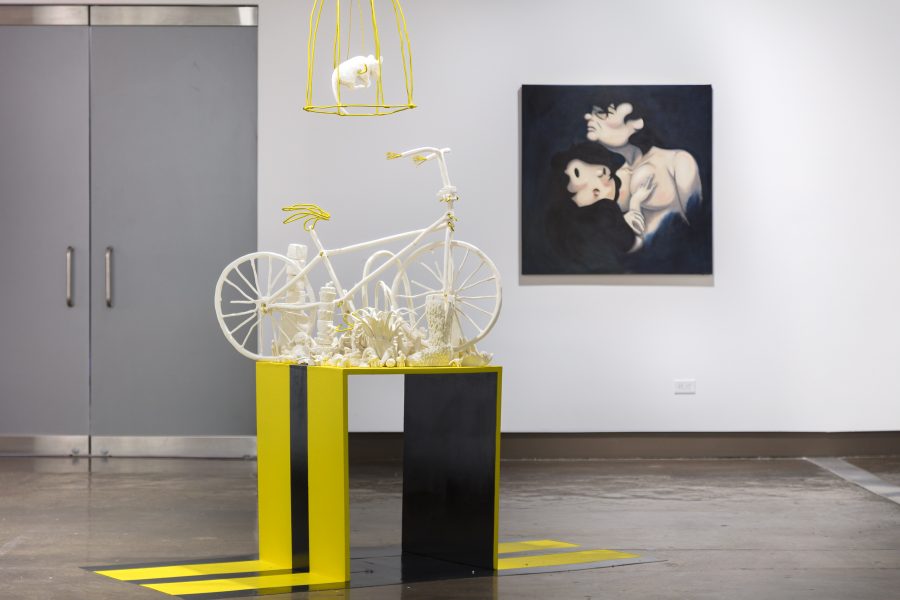 Installation view of an exhibition with a large cartoonish portrait painting in the background. Black and yellow pedestal with a bicycle is in the foreground. A white rat in a yellow cage is suspended above the bicycle.