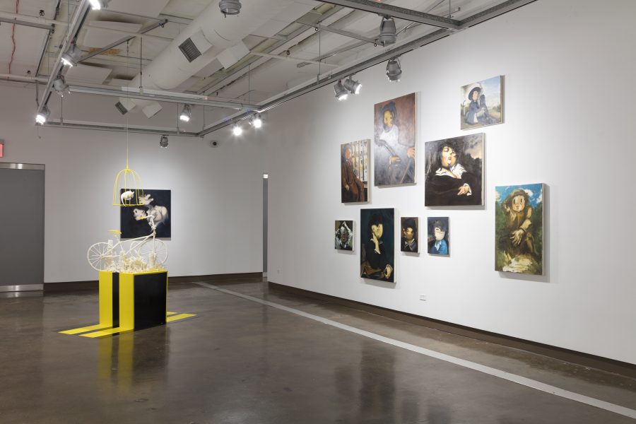 Installation view of exhibition that consists of salon style hanging of cartoonish portraiture, and a black and yellow pedestal with a small bicycle that rests on top.